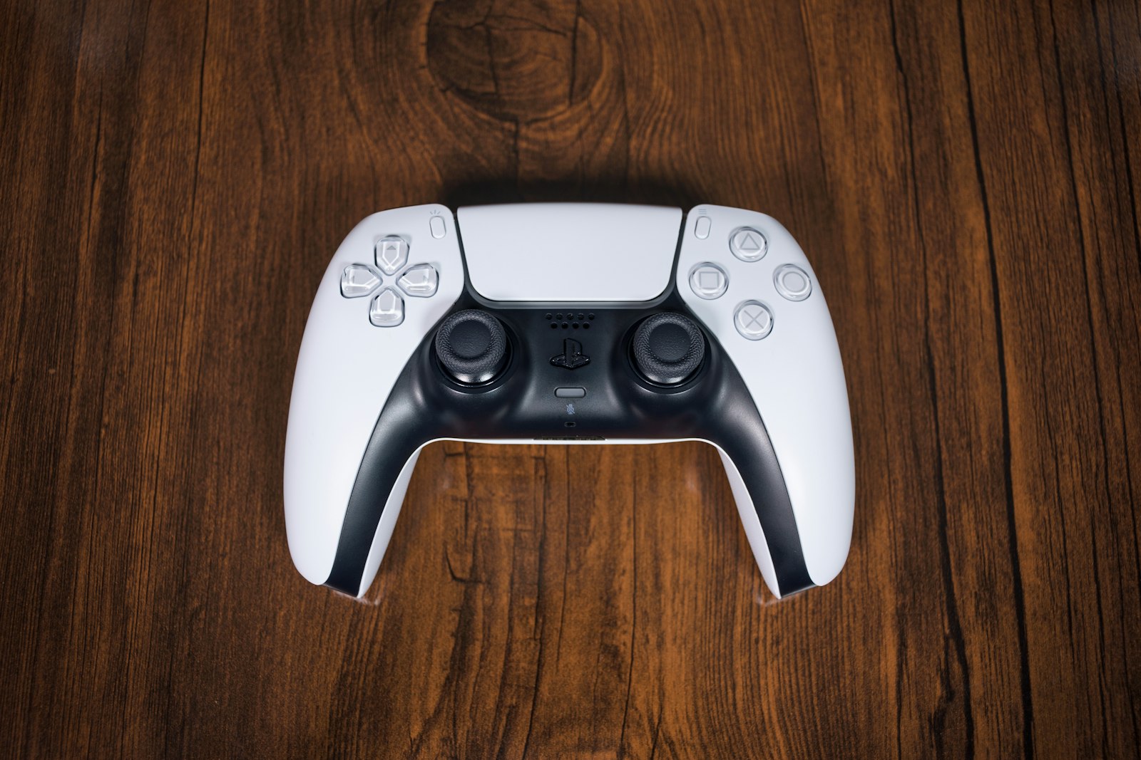 White PlayStation 5 DualSense controller lying face-up on a dark wooden desk.